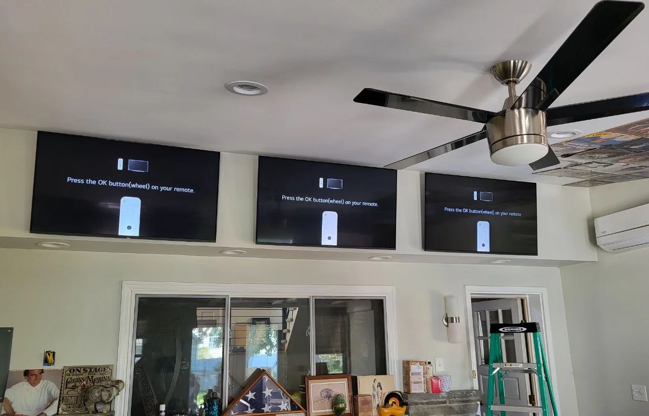 Three televisions mounted above windows beneath a ceiling fan, with a ladder in the room.