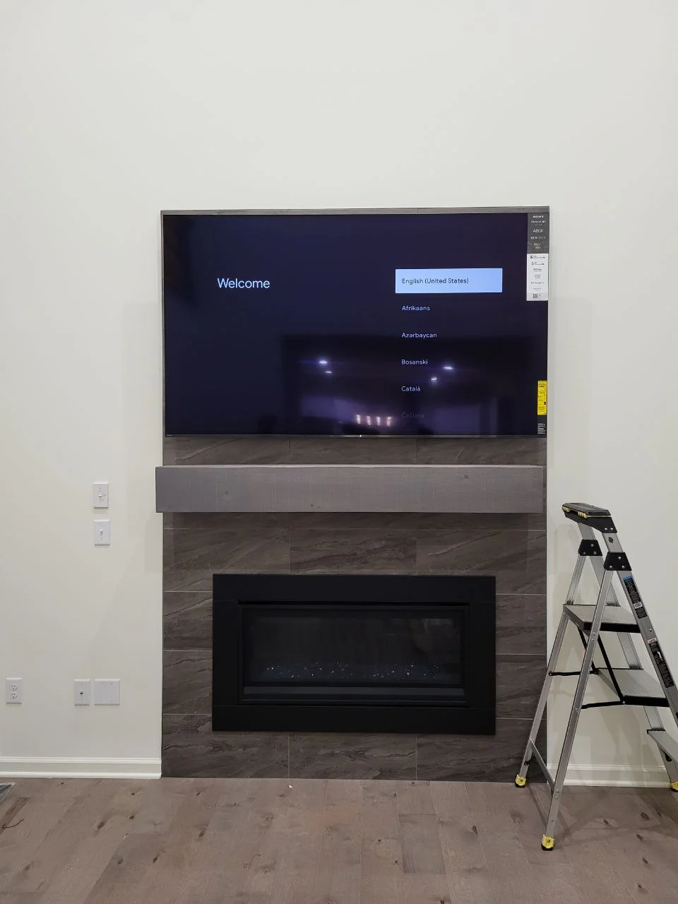 Television displaying a setup screen above a gray fireplace surround, with a ladder beside it.